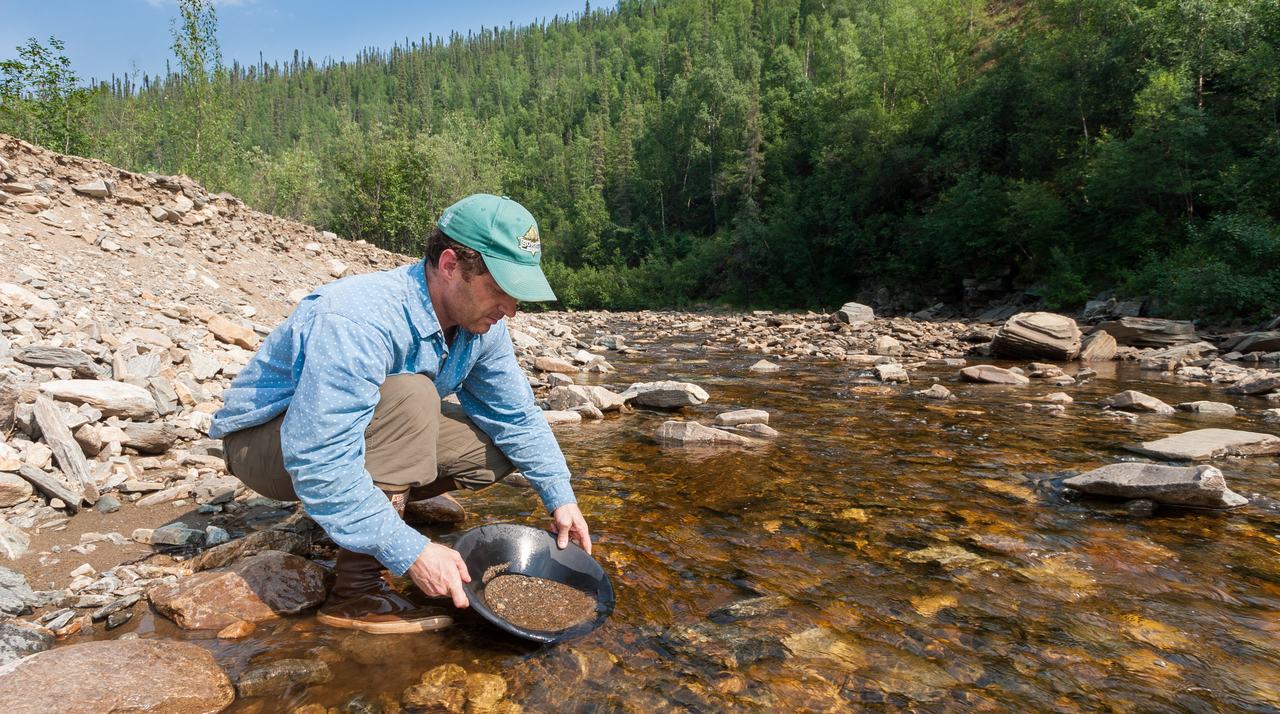 A prospector crouched at the water's edge, panning river gravel down toward the concentrate.