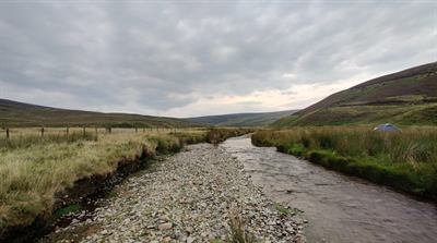 A shallow burn beside a wide bar of grey stones, with heather-covered hills under a cloudy sky