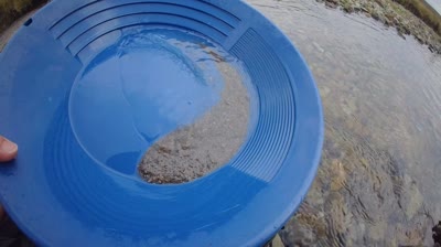 A blue plastic gold pan held over a clear, shallow stream