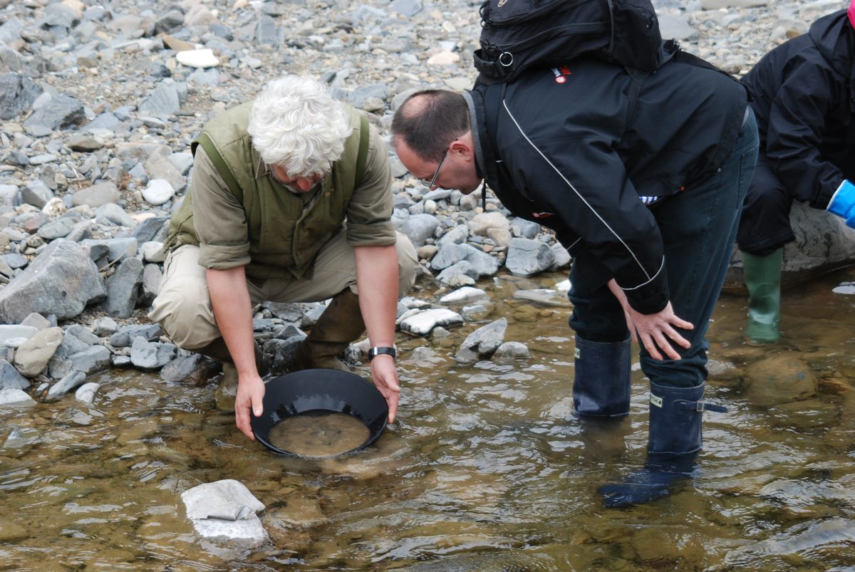 Vincent Thurkettle crouching at the water's edge with a gold pan while two people in waterproofs lean in to watch.